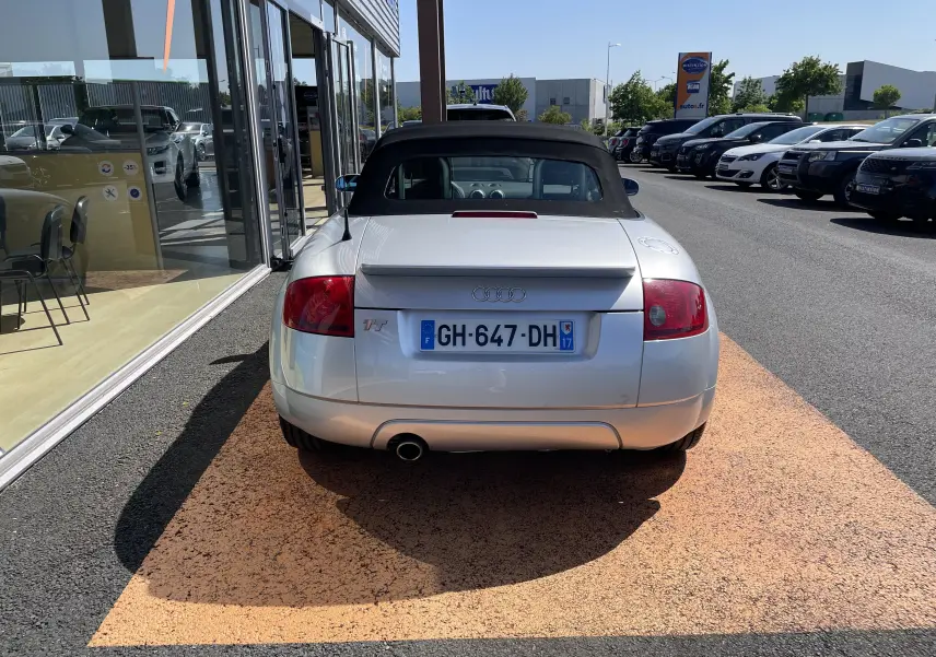 Vue arrière d'une Audi TT Roadster gris argent de 2002 avec capote noire et aileron intégré, stationnée devant un showroom.