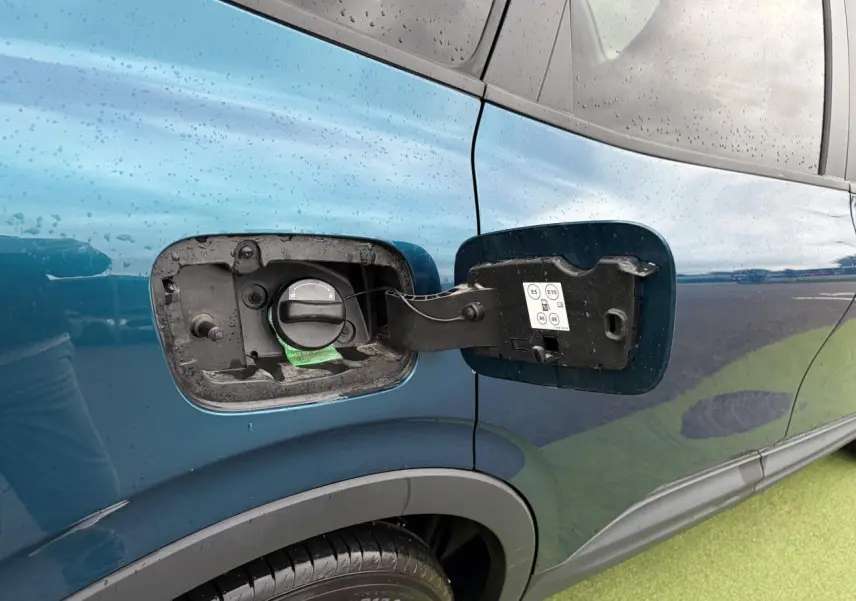 Gros plan sur la trappe à carburant ouverte côté gauche d’un Renault Arkana bleu Zanzibar métal sous la pluie.