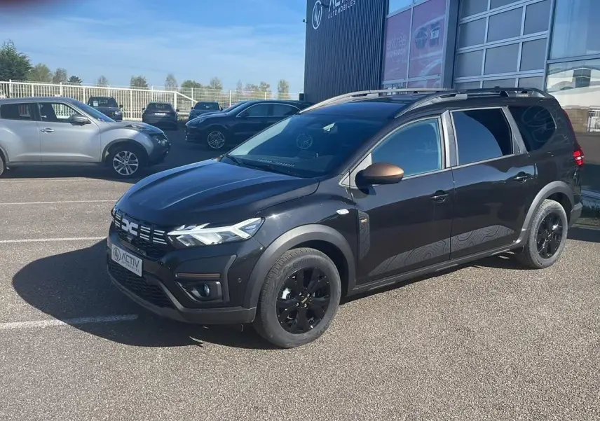 Dacia Jogger noir nacré en 3/4 avant droit avec jantes noires et barres de toit visibles sur parking.