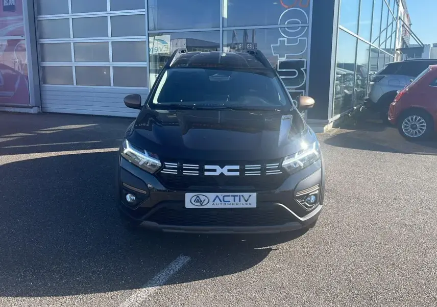 Vue de face du Dacia Jogger noir nacré avec calandre distinctive et phares LED allumés, stationné devant un garage.