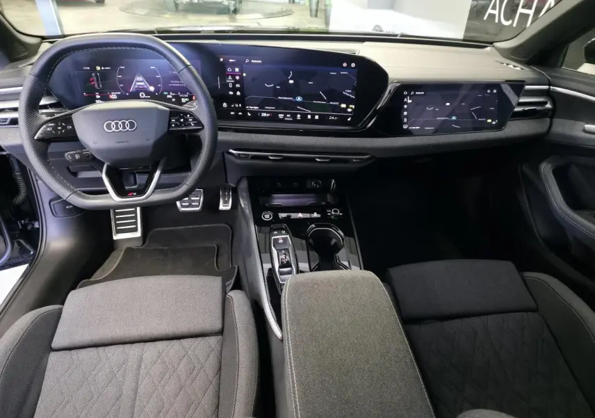 Vue intérieure avant de l'Audi A5 2025 S line, tableau de bord noir avec double écran numérique et volant sport cuir.