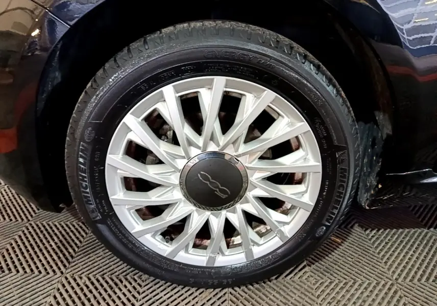 Gros plan sur la roue avant droite d'une FIAT 500 noire, mettant en valeur la jante argentée au design étoilé.