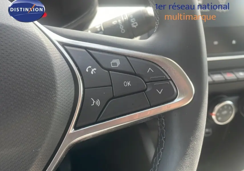 Gros plan sur les commandes multifonctions à droite du volant noir d’une Renault Clio blanche, avec tableau de bord en arrière-plan.