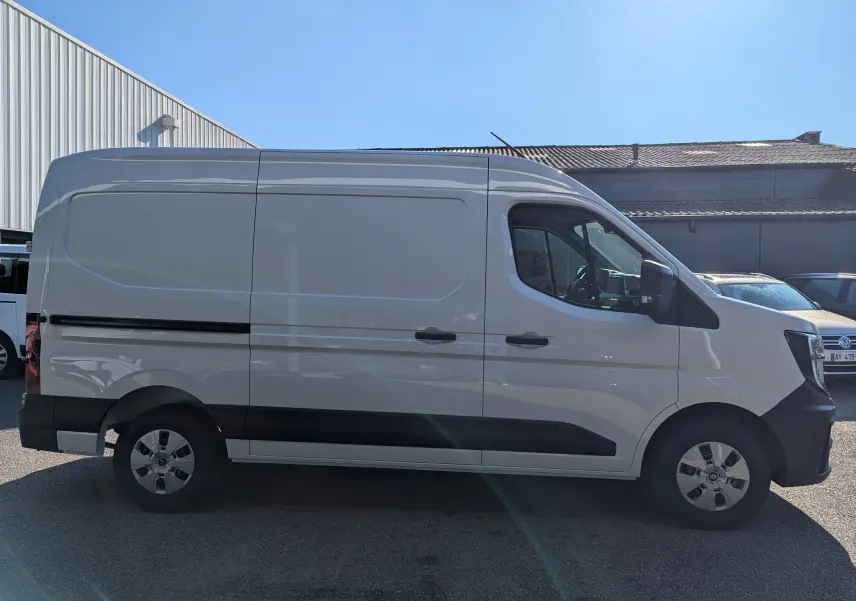 Vue latérale droite d'un utilitaire Renault Master blanc L2H2 avec jantes acier et protections noires sur fond de bâtiment industriel.