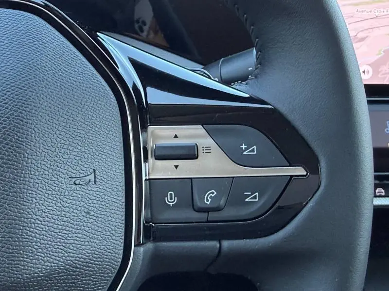 Gros plan sur les commandes multifonctions à gauche du volant noir de la Peugeot 308 hybride 2025.