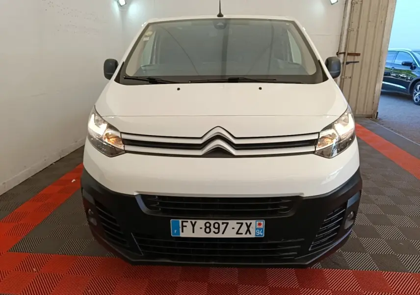 Vue frontale d'un Citroën Jumpy XS blanc avec pare-chocs noir et phares allumés en intérieur.