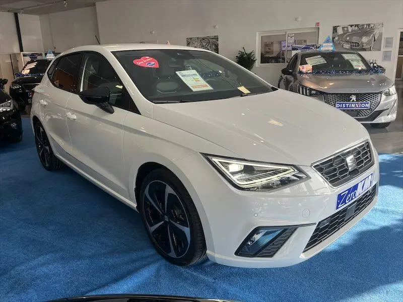 SEAT Ibiza 2024 blanc Nevada vue 3/4 avant droit en showroom avec jantes noires et phares LED allumés.
