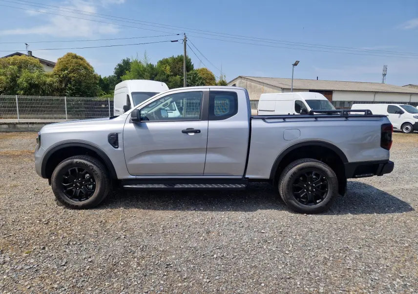 Vue de profil côté gauche du Ford Ranger Super Cabine gris Iconic avec marchepied et jantes noires sur terrain gravelé.
