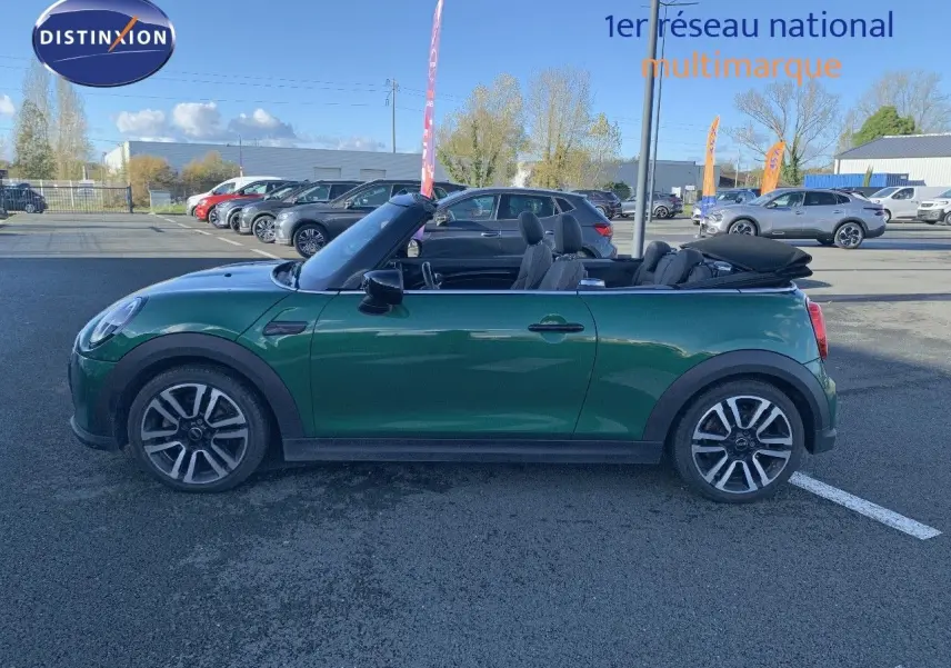 Profil côté gauche d'une MINI Cabriolet F57 2023 British Racing Green avec capote ouverte sur parking.