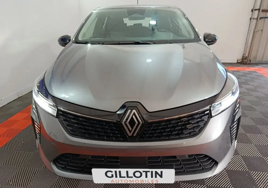 Vue avant rapprochée d'une Renault Clio gris clair avec calandre noire et phares LED allumés dans un garage.