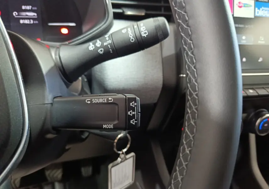 Gros plan sur le volant cuir synthétique et les commandes au volant d’une Renault Clio gris clair, tableau de bord partiellement visible.