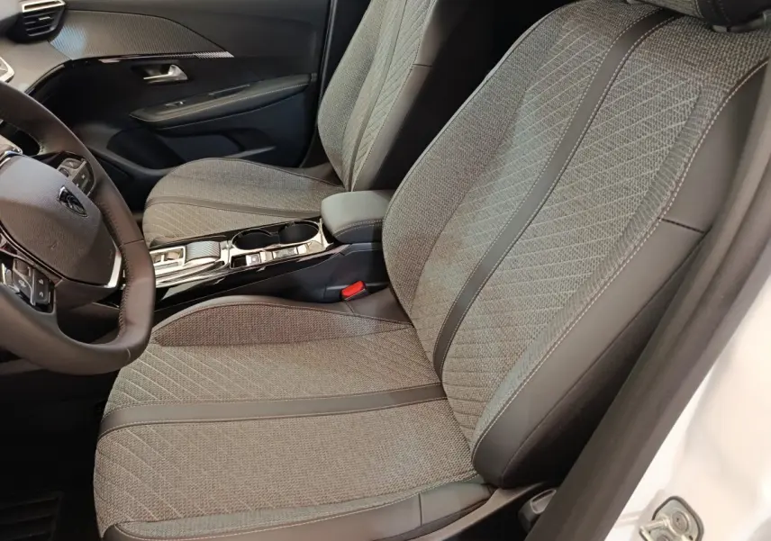 Vue intérieure côté conducteur de la Peugeot 208 électrique blanche 2024, sièges tissu gris avec surpiqûres et volant cuir.