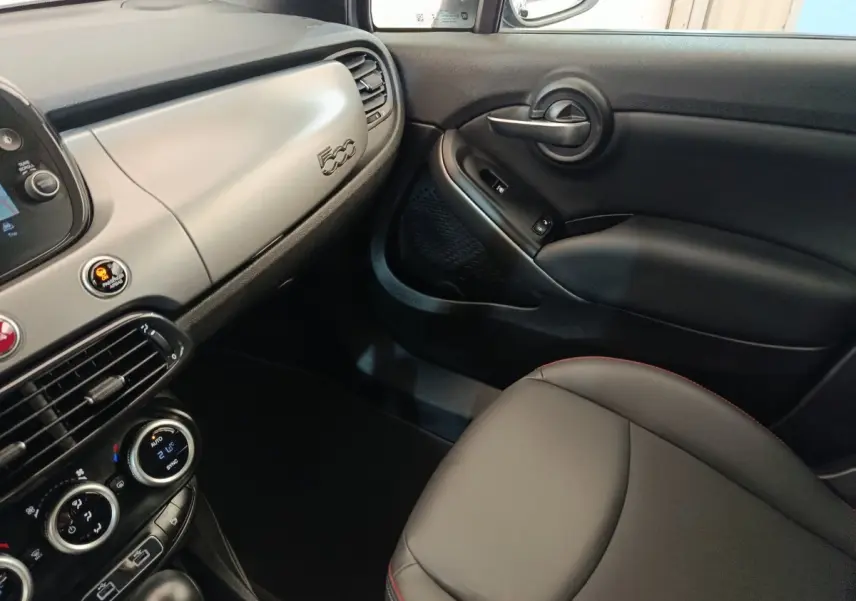 Intérieur côté passager de la Fiat 500X 2021, tableau de bord gris clair avec logo 500 et sellerie noire avec surpiqûres rouges.