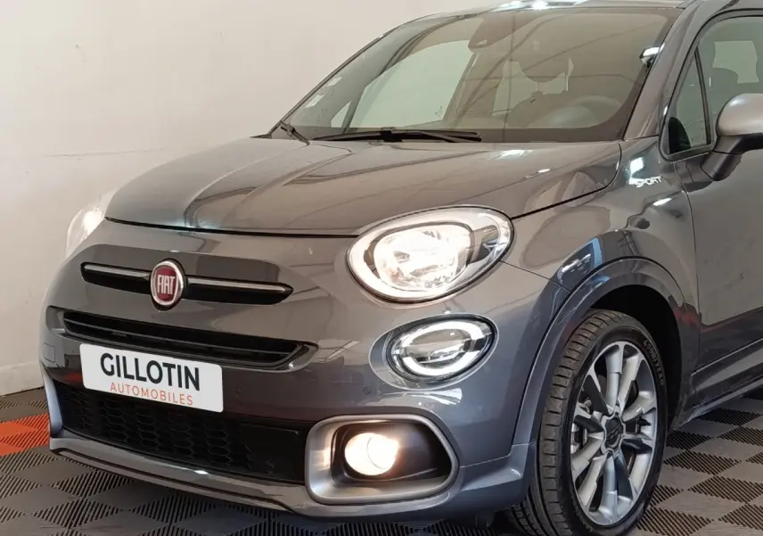 Vue 3/4 avant droite d'une FIAT 500X gris foncé avec phares allumés et badge Sport sur l'aile.