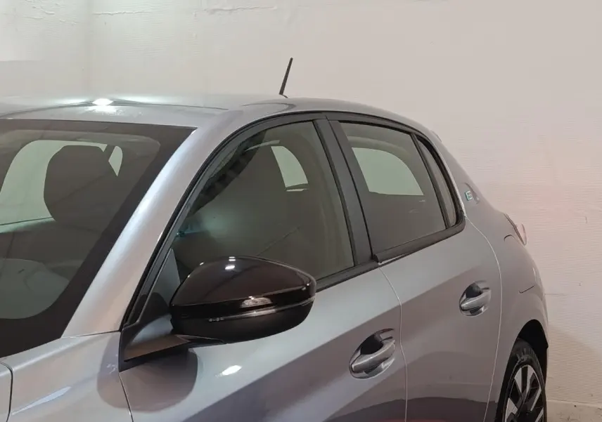 Vue côté gauche avant d'une Peugeot 208 électrique gris clair avec rétroviseur noir brillant et poignées de porte assorties.