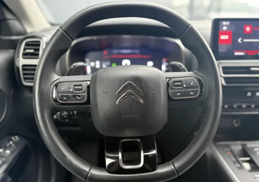 Vue rapprochée du volant en cuir noir du Citroën C5 Aircross 2024 avec commandes intégrées et tableau de bord numérique flou.
