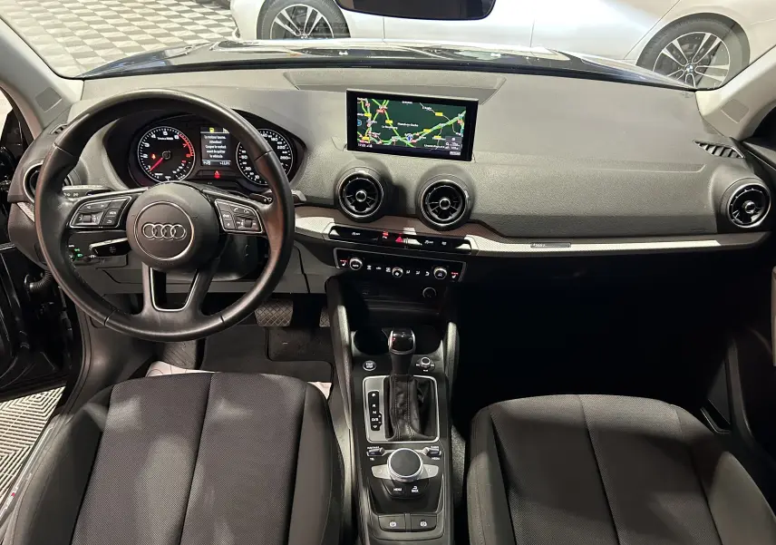 Coffre spacieux et propre de l'Audi Q2 noir métal, vue intérieure depuis l'ouverture arrière, avec tapis de sol gris foncé.