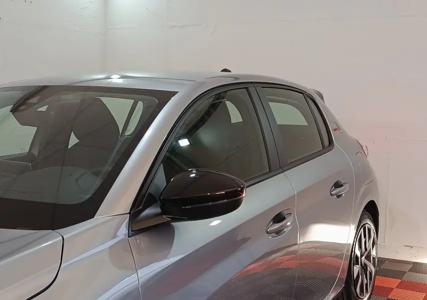 Vue de côté droit avant d'une Peugeot 208 Hybrid gris clair avec rétroviseur noir et jantes distinctives.