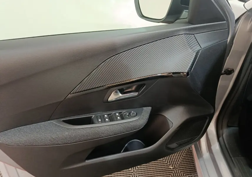 Vue intérieure du panneau de porte côté conducteur gris clair de la Peugeot 208 Hybrid 2025 avec commandes électriques et insert effet carbone.