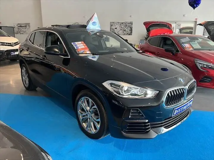 BMW X2 noir métal en 3/4 avant droit, avec toit ouvrant, exposée en showroom sur sol bleu.