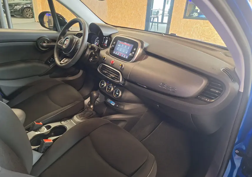 Intérieur de la FIAT 500X 2024 bleu, vue côté conducteur, tableau de bord avec écran tactile et volant multifonctions.
