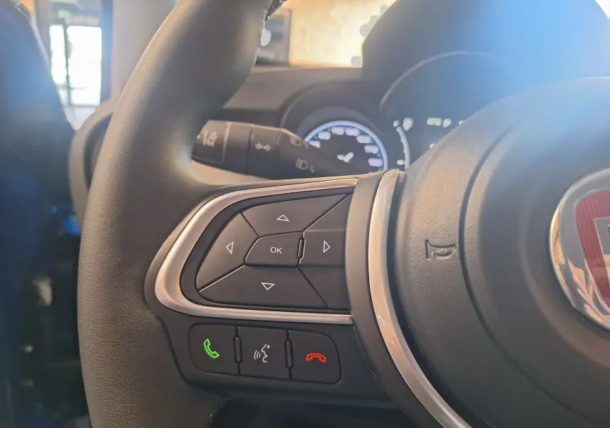 Gros plan sur les commandes du volant à gauche d'une FIAT 500X bleu 2024 avec logo rouge central visible.