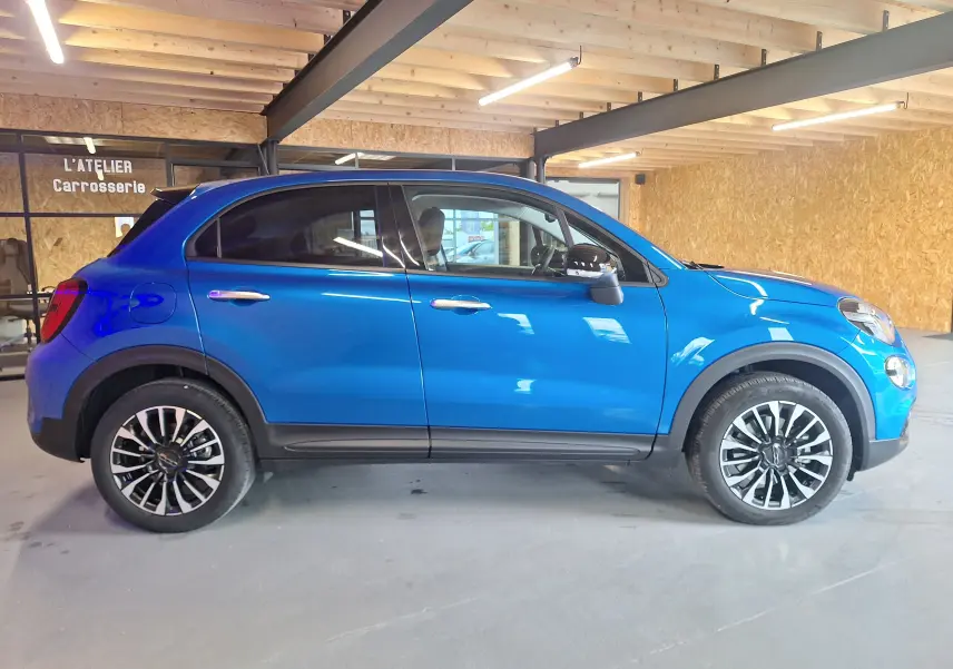 Profil latéral droit d'un FIAT 500X bleu métallisé 2024 avec jantes alliage et protections noires sur châssis.