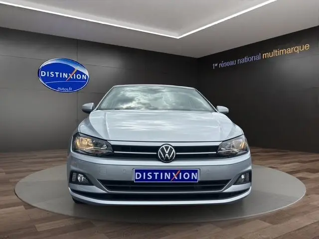 Volkswagen Polo VI Lounge Business gris vue de face dans un showroom avec phares allumés et logo Distinxion visible.