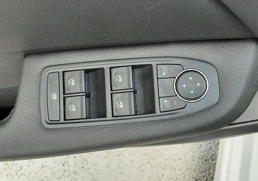 Gros plan sur les commandes de lève-vitres et réglage des rétroviseurs côté conducteur d’une Renault Clio V blanche.