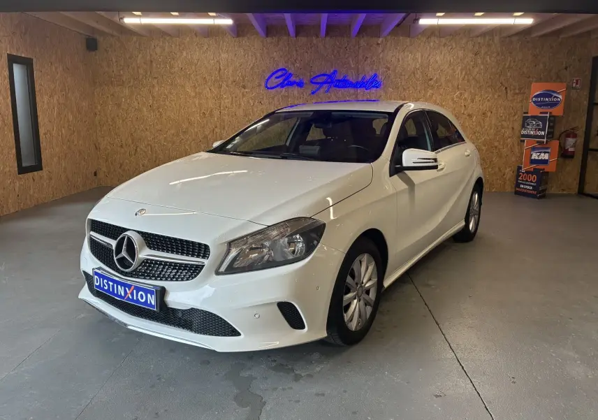 Mercedes Classe A 200D blanche vue de 3/4 avant droit dans un showroom avec mur en bois et enseigne bleue lumineuse.