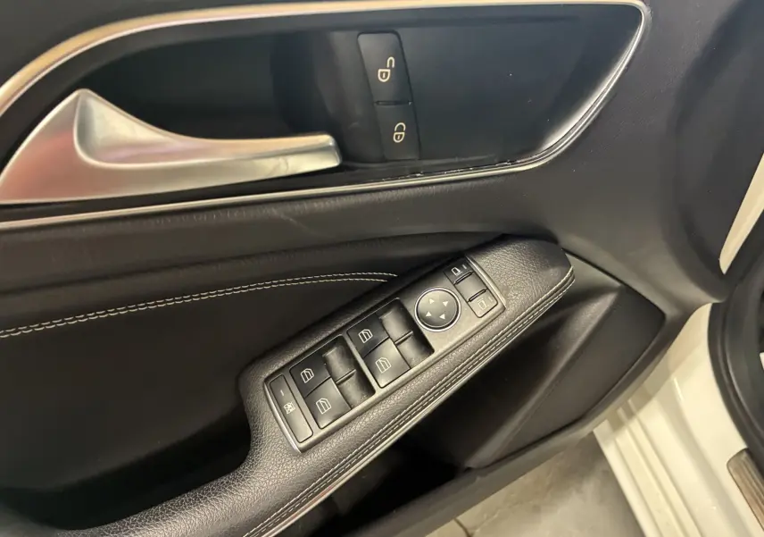 Gros plan sur la porte côté conducteur d'une Mercedes Classe A blanche, montrant les commandes de vitres et la poignée intérieure argentée.