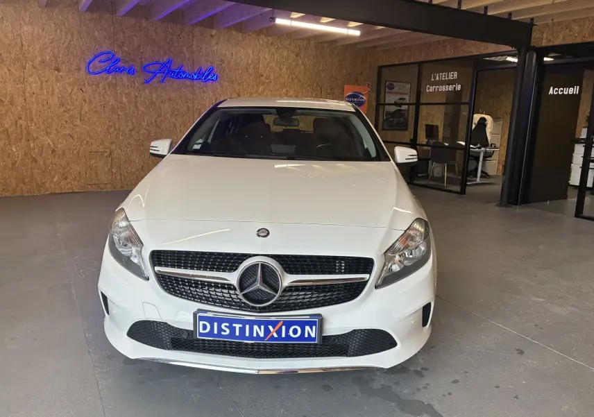 Vue frontale d'une Mercedes Classe A 200D blanche dans un showroom avec un mur en bois et enseigne néon bleue.