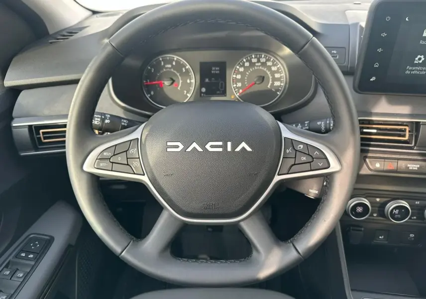 Vue intérieure centrée sur le volant noir avec logo Dacia, tableau de bord analogique et écran tactile à droite.