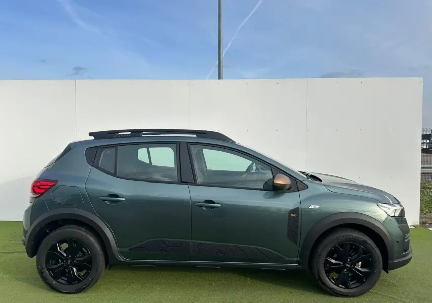 Vue de profil côté gauche d'une Dacia Sandero III Stepway vert cèdre avec jantes noires et barres de toit.