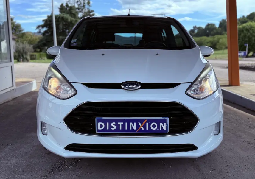 Vue frontale d'une Ford B-MAX blanche avec phares allumés et plaque d'immatriculation Distinxion visible.