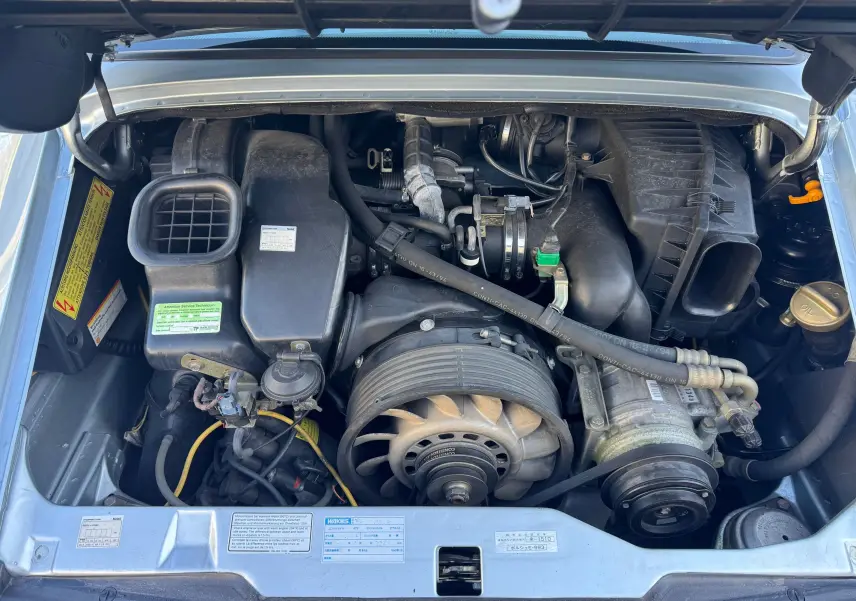 Vue plongeante sur le moteur essence d'une Porsche 911 Carrera 2 gris polaire de 1996, capot ouvert.