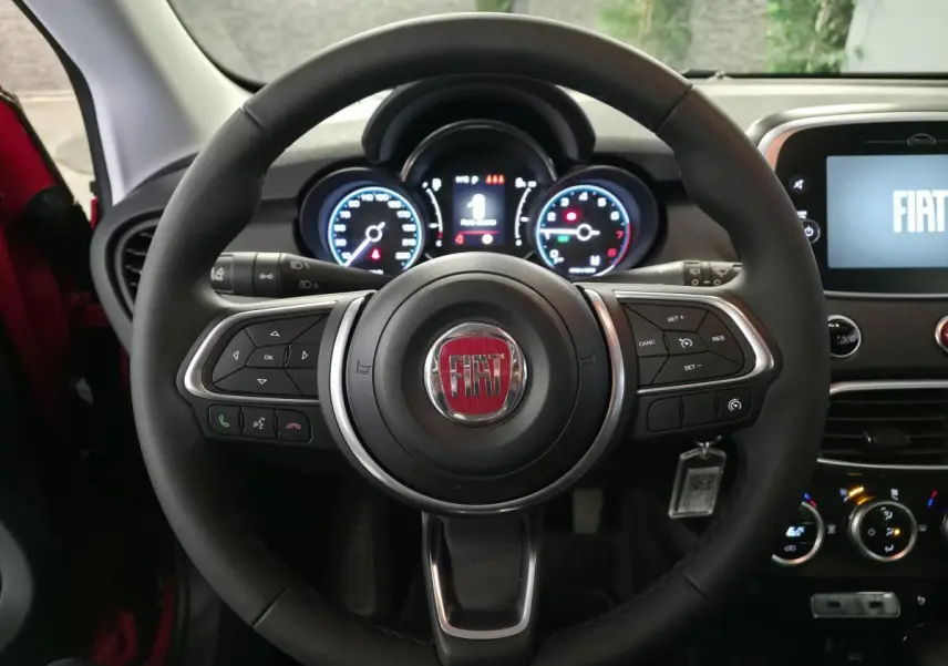Vue intérieure centrée sur le volant noir soft-touch avec logo FIAT rouge et tableau de bord éclairé du 500X rouge.
