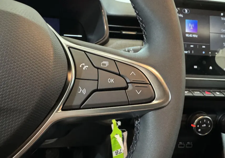 Gros plan sur les commandes au volant côté gauche d’une Renault Clio gris clair, avec écran tactile en arrière-plan.