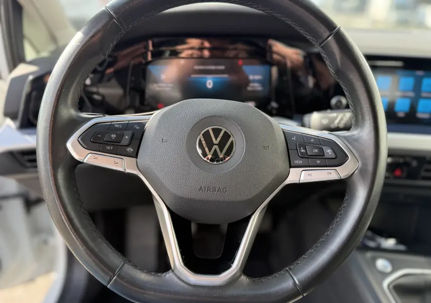 Vue rapprochée du volant en cuir noir avec logo Volkswagen, tableau de bord digital et écran tactile en arrière-plan d'une Golf blanche.