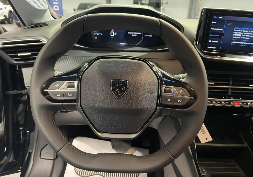 Vue rapprochée du volant cuir noir de la Peugeot 208 1.2i Hybrid 2025 avec tableau de bord numérique et écran tactile.
