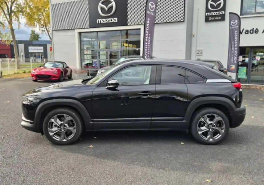 Vue de profil côté gauche d'un Mazda MX-30 électrique noir devant un concessionnaire Mazda avec logos visibles.