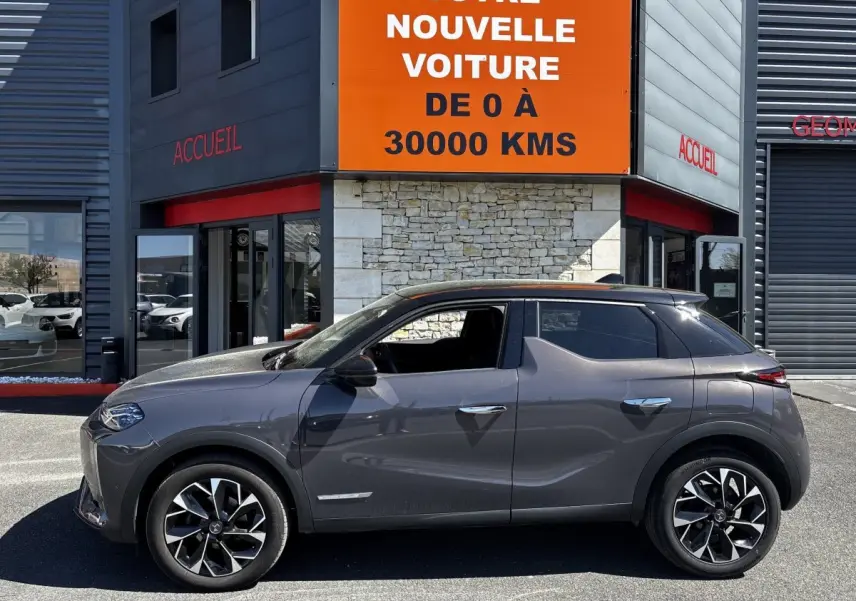 Vue de profil côté gauche d'une DS3 1.2 PureTech 130 gris foncé garée devant un bâtiment avec panneau orange.