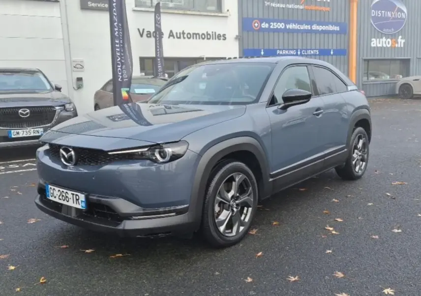 Vue 3/4 avant d'un Mazda MX-30 gris électrique, avec jantes distinctives et protections noires autour des passages de roues.