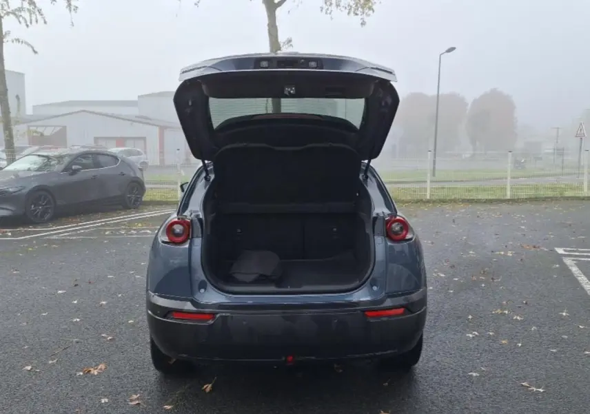 Vue arrière de la Mazda MX-30 gris 2021 avec coffre ouvert, montrant l'espace de chargement vide sur un parking extérieur.