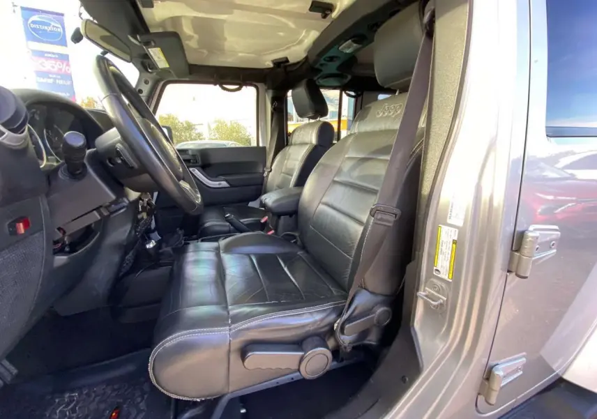 Vue intérieure côté conducteur du Jeep Wrangler gris clair 2012, mettant en valeur les sièges en cuir noir avec logo Jeep brodé.