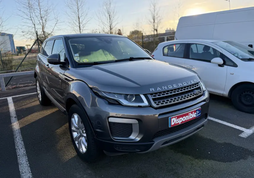 Range Rover Evoque gris vue 3/4 avant droit, avec calandre distinctive et plaque "Disponible" sur parking extérieur.