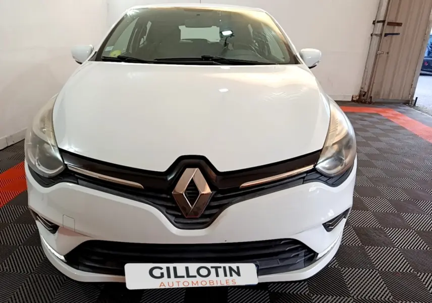 Vue frontale d'une Renault Clio blanche 2016 avec logo Renault proéminent et phares effilés dans un garage.