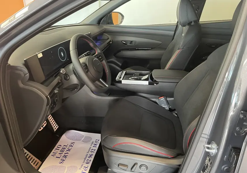 Intérieur du Hyundai Tucson 2025 gris Ecotronic, vue côté gauche, sièges noirs avec surpiqûres rouges et tableau de bord numérique.