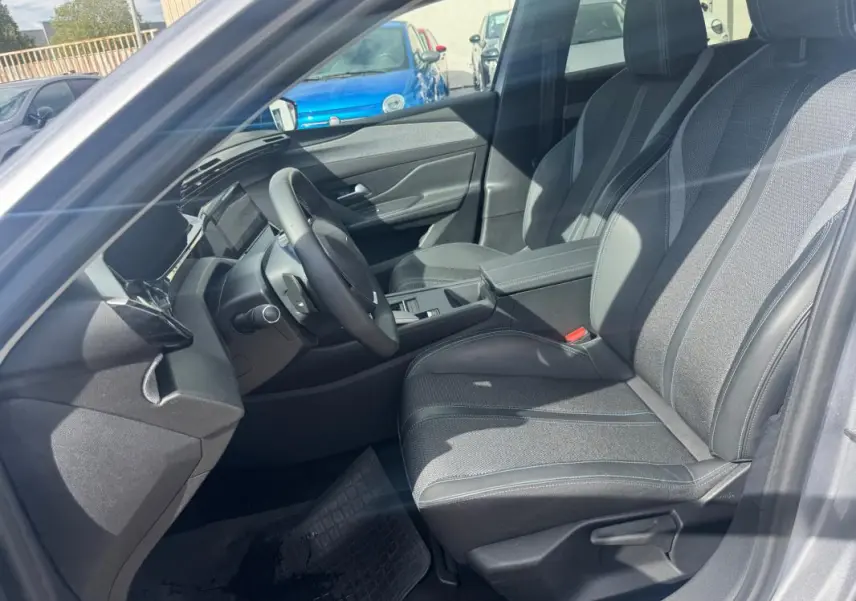 Vue intérieure côté conducteur de la Peugeot 308 Hybrid 145 gris, avec sièges tissu et accoudoir central visible.