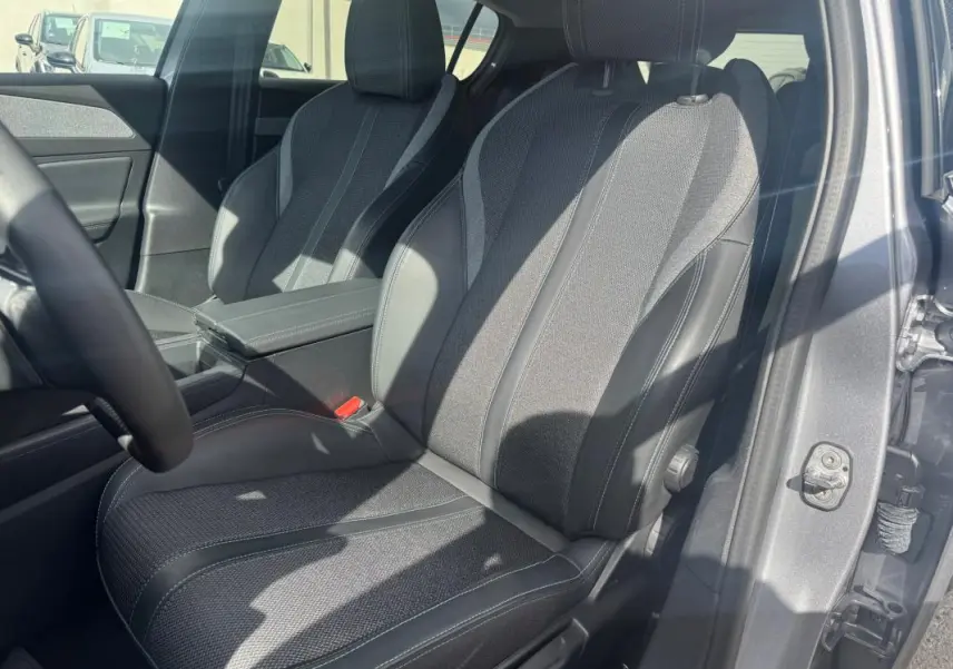 Vue intérieure côté conducteur d’un Peugeot 308 gris 2025, sièges tissu et cuir avec accoudoir central visible.